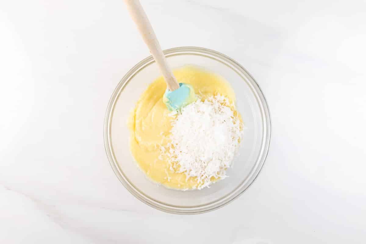 A glass bowl containing yellow cake batter and a pile of shredded coconut, with a blue silicone spatula resting inside, on a white surface.