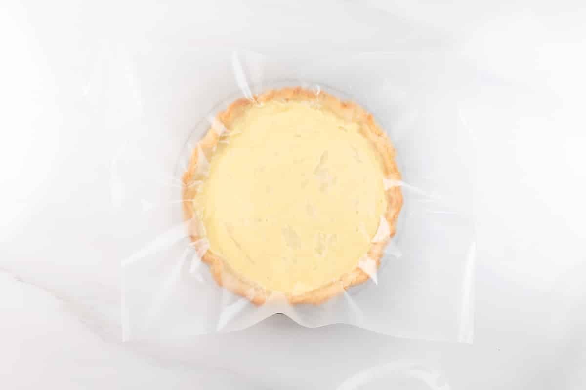 A round pie with a creamy yellow filling sits on a white surface, partially covered by a sheet of transparent plastic wrap.