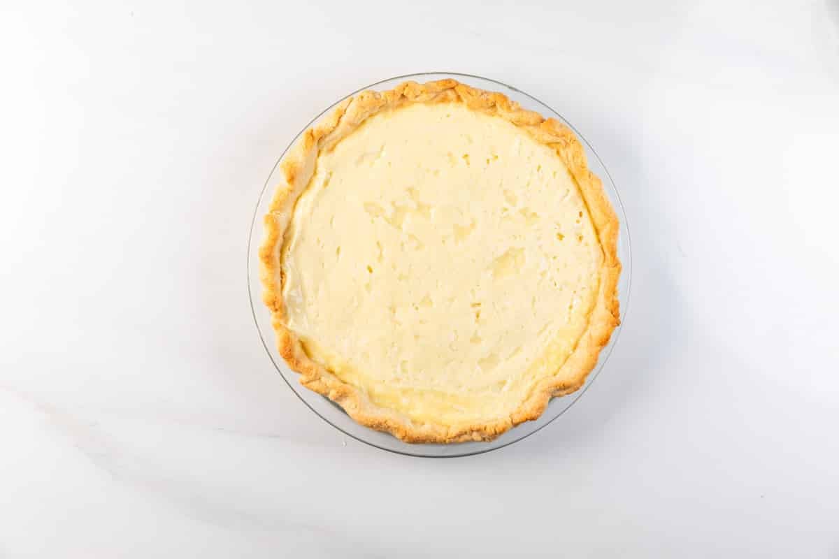 A whole baked pie with a golden crust and creamy, pale filling sits on a white plate against a white marble background.