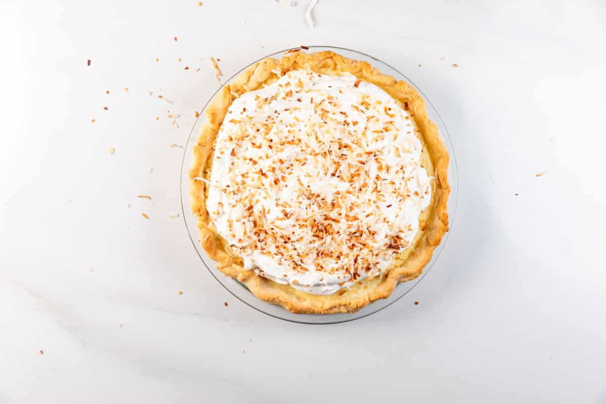 A coconut cream pie topped with whipped cream and toasted coconut flakes sits on a clear glass pie dish on a white surface. Some toasted coconut flakes are scattered around the pie.
