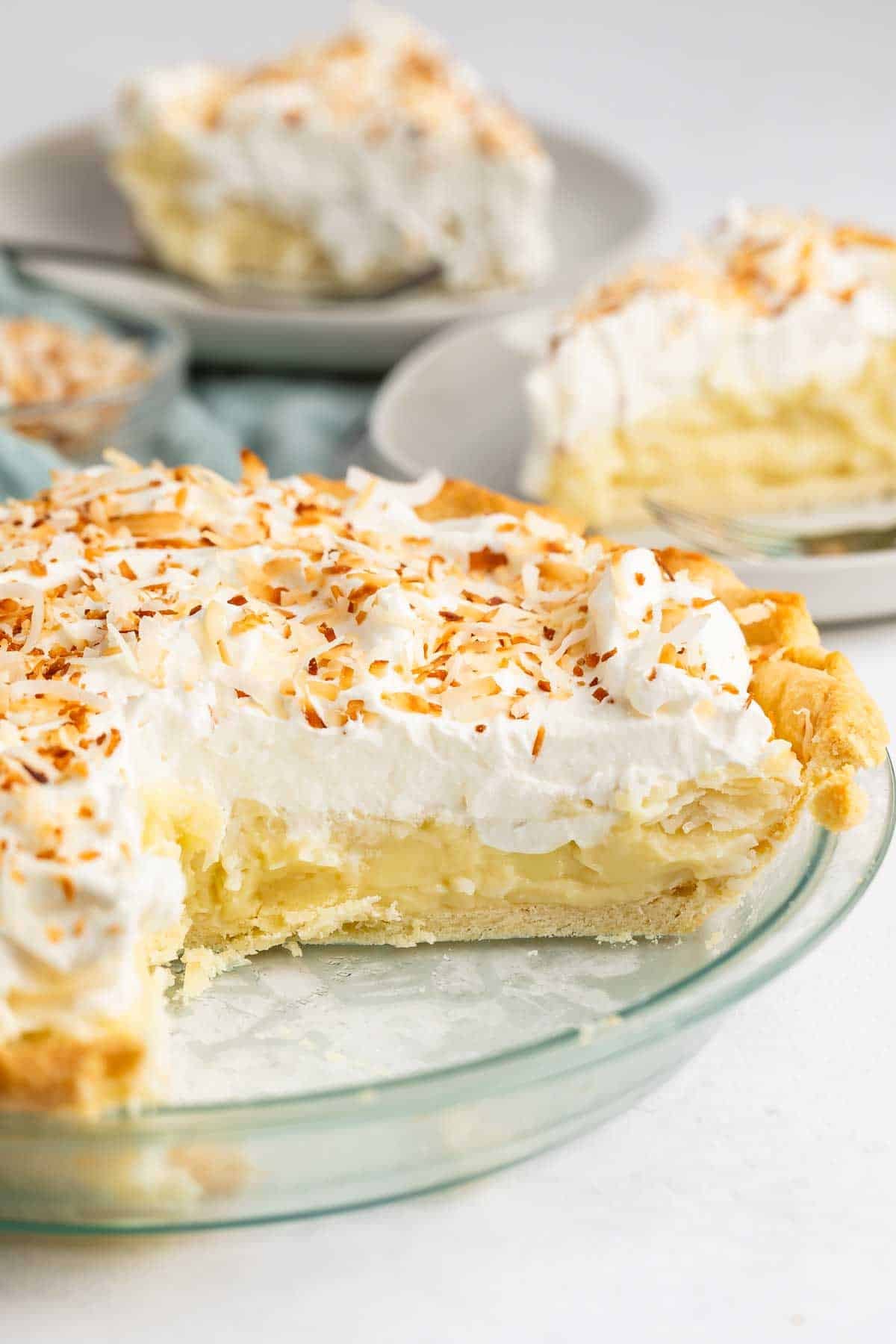 A glass pie dish filled with coconut cream pie, topped with whipped cream and toasted coconut flakes. A slice is missing, and two plated slices of coconut cream pie are visible in the background.