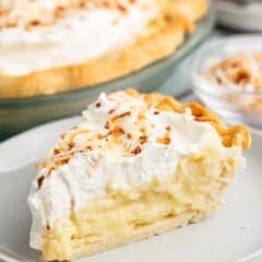 A slice of coconut cream pie topped with whipped cream and toasted coconut flakes sits on a plate, while the rest of the coconut cream pie and a small bowl of toasted coconut are visible in the background.