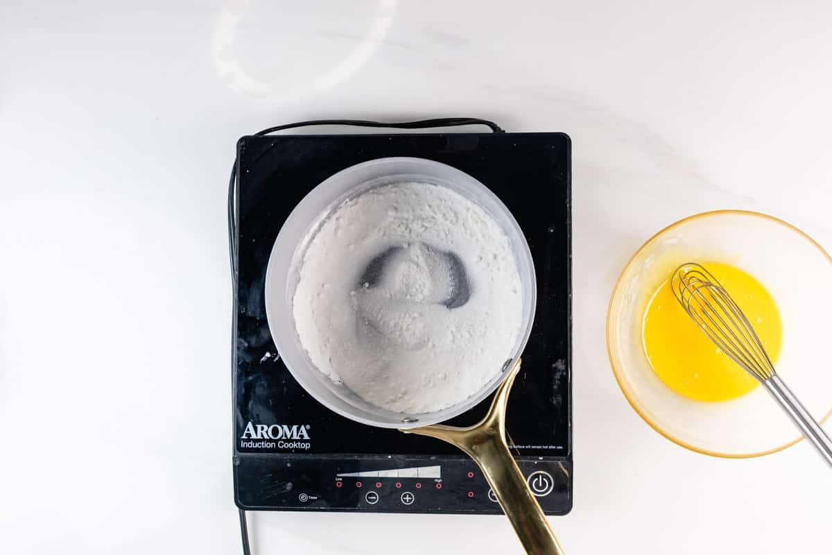 A white saucepan with milk heating on an induction cooktop sits next to a bowl of whisked eggs on a white surface, with a gold-handled whisk resting nearby.