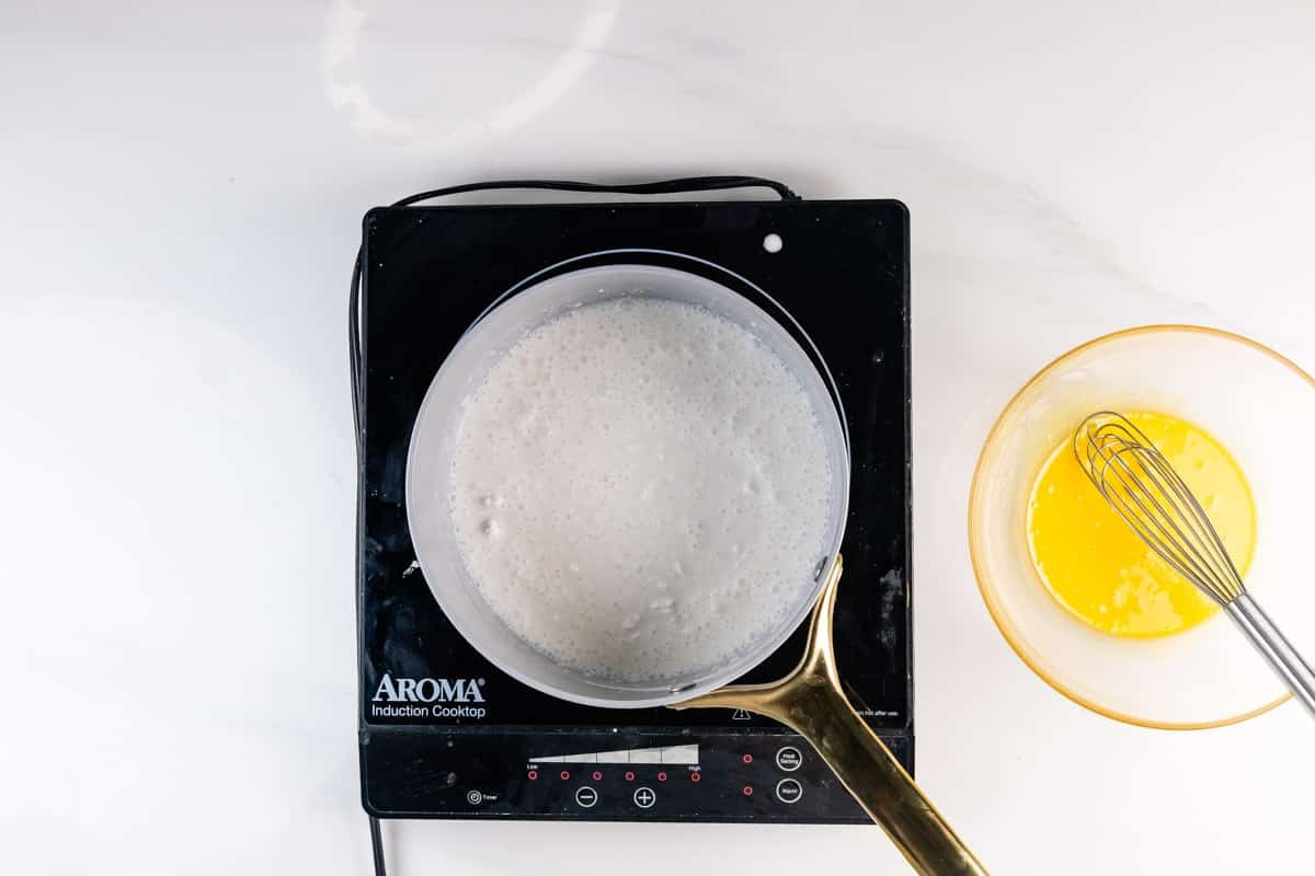 A saucepan with a white bubbling mixture sits on an Aroma induction cooktop. Next to it is a bowl of melted yellow liquid with a metal whisk inside, all on a white countertop.