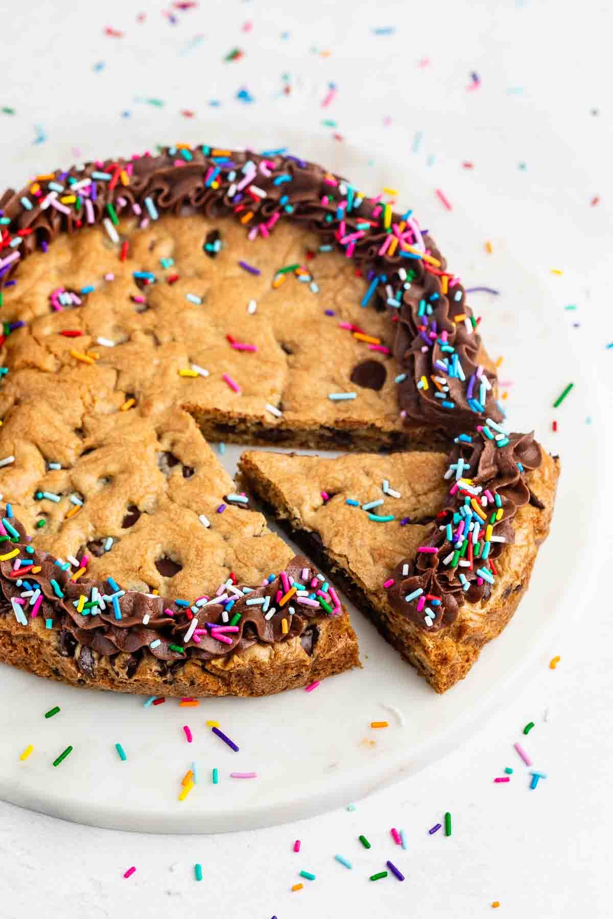 A large chocolate chip cookie cake decorated with chocolate frosting and colorful sprinkles sits on a white plate. One slice is partly removed, with sprinkles scattered around the plate.