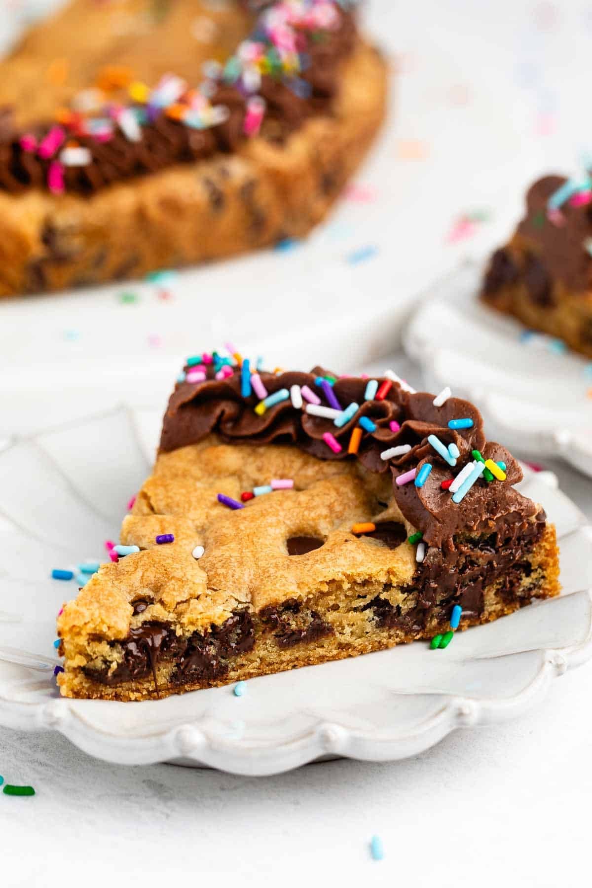 A slice of chocolate chip cookie cake with chocolate frosting and colorful sprinkles sits on a white plate, with the rest of the cookie cake and another slice in the background.