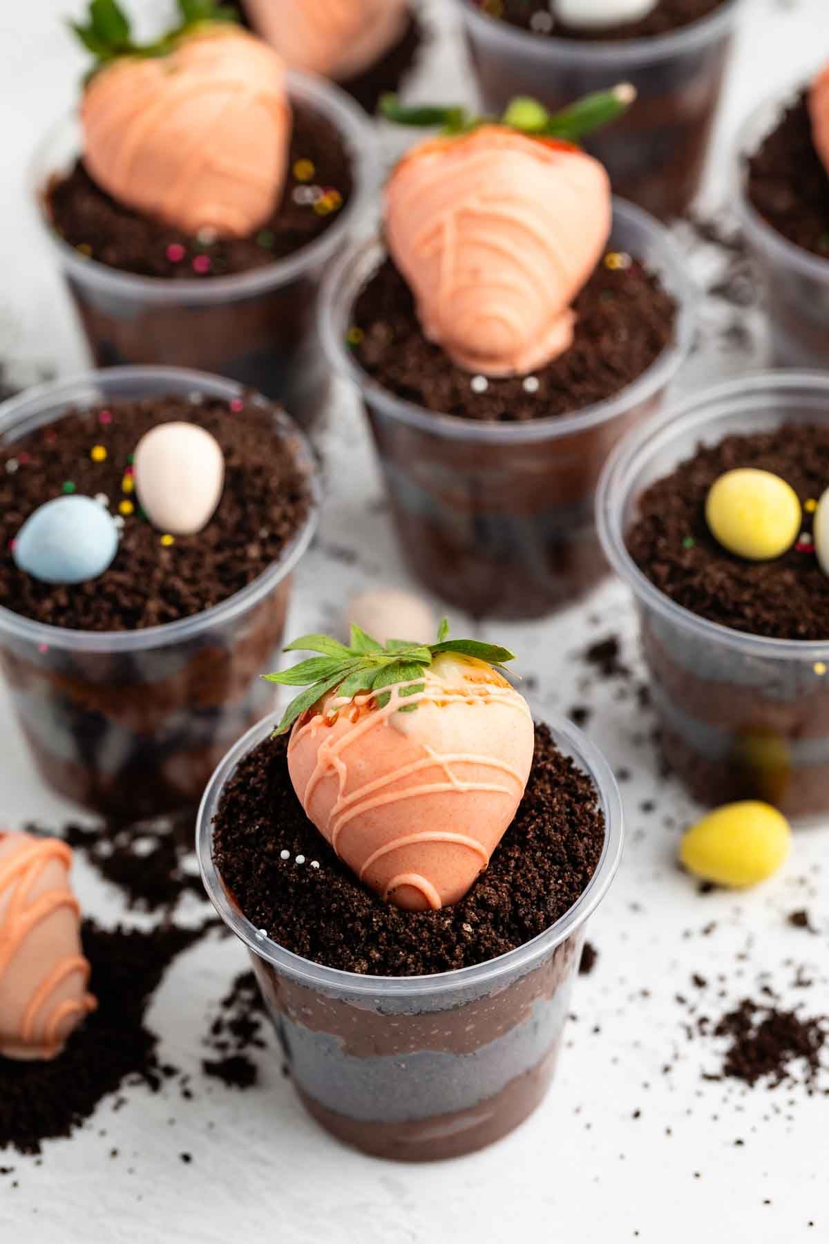 Clear plastic cups filled with layered chocolate pudding and crushed cookies, each topped with a strawberry coated in orange chocolate to resemble a carrot, and some decorated with small candy eggs.