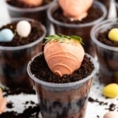 A clear plastic cup filled with chocolate pudding and crushed cookies, topped with a strawberry coated in orange chocolate. More cups with similar toppings are in the background, along with small pastel candy eggs.