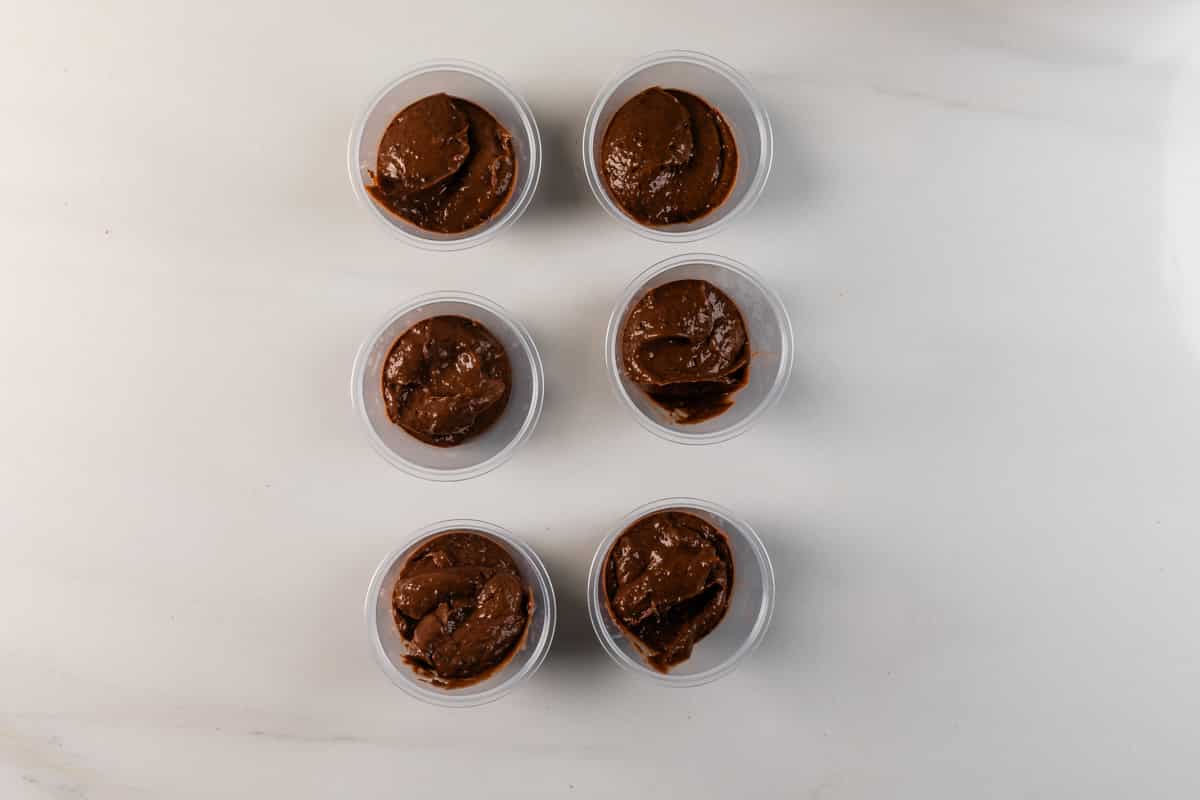 Six small clear cups filled with chocolate mousse are arranged neatly in two vertical columns on a white marble surface.