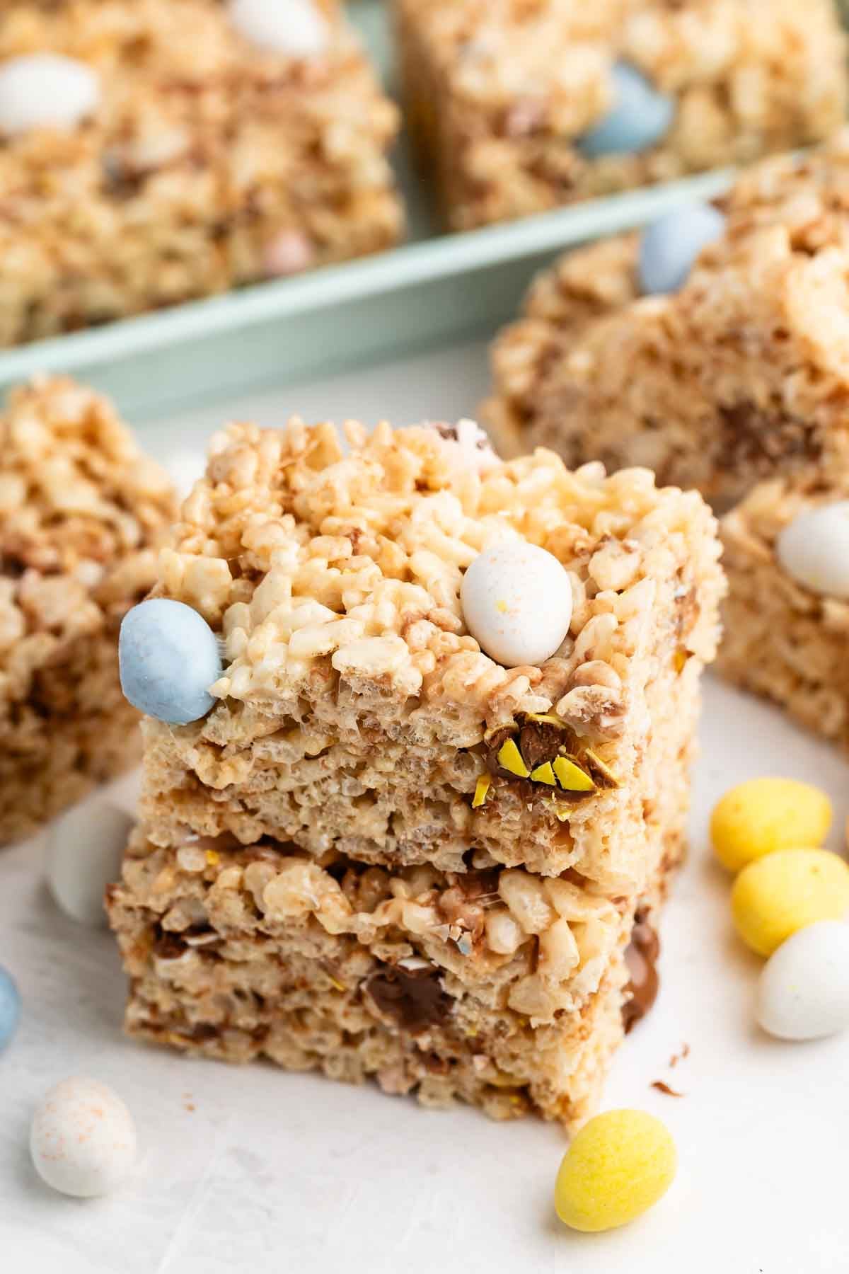 Two rice krispie treat squares stacked on top of each other, with small pastel-colored candy eggs mixed in and scattered around, and more treats visible in the background.
