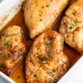 Four cooked chicken breasts in a white baking dish, covered in a brown sauce and garnished with chopped herbs.