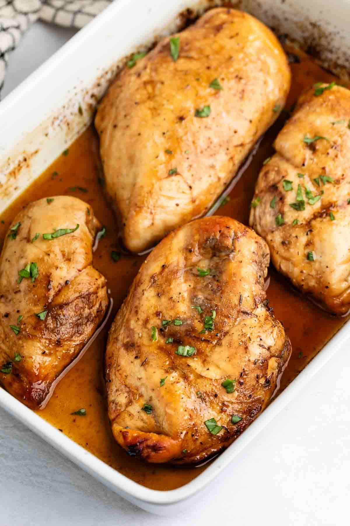 Four cooked chicken breasts in a white baking dish, covered in a brown sauce and garnished with chopped herbs.