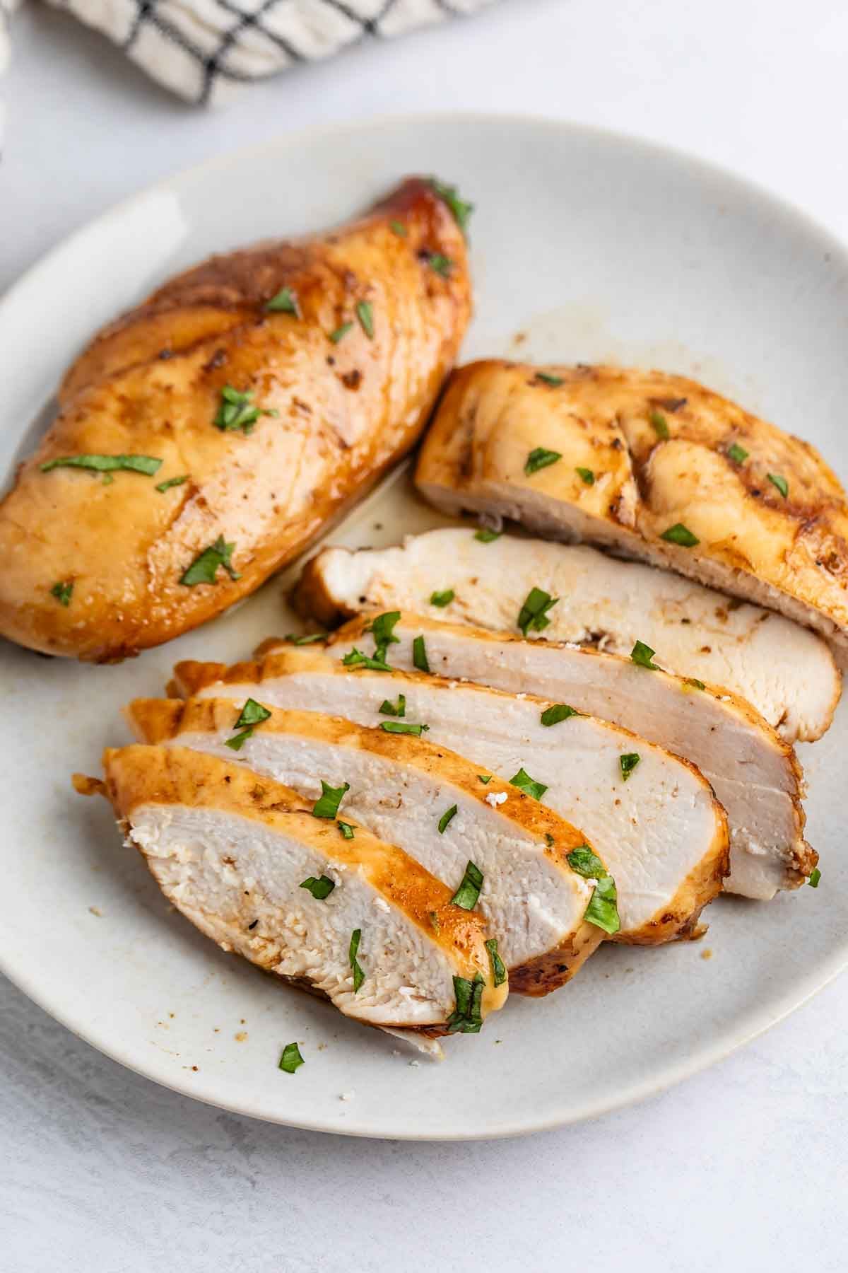 A plate with a whole and sliced cooked chicken breast, garnished with chopped herbs, on a light-colored surface.