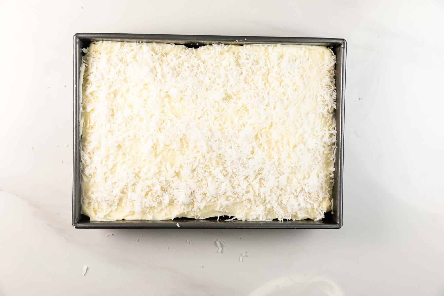 A square baking pan filled with a dessert topped with a thick layer of white shredded coconut on a smooth white surface.