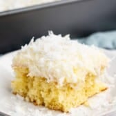 A slice of yellow cake topped with white frosting and shredded coconut sits on a white plate, with a baking pan of the same cake in the background.