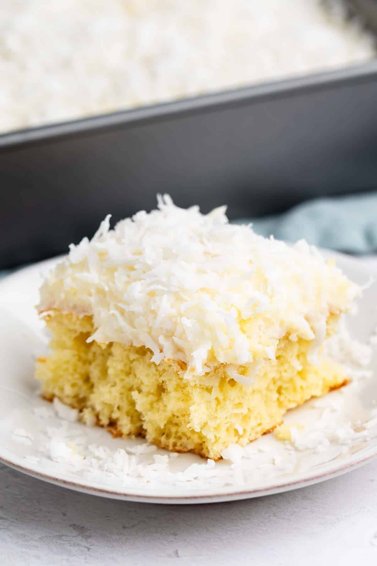 A slice of yellow cake topped with white frosting and shredded coconut sits on a white plate, with a baking pan of the same cake in the background.