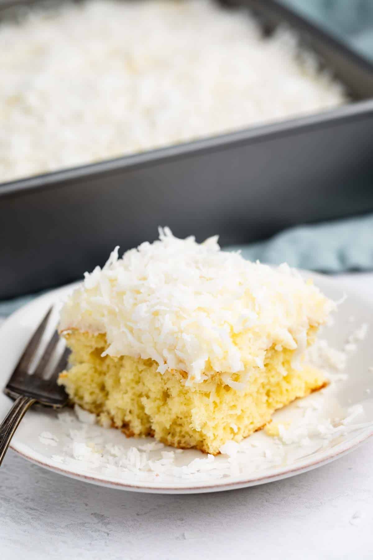 A slice of yellow cake topped with shredded coconut sits on a white plate with a fork, with a larger pan of the same cake in the background.