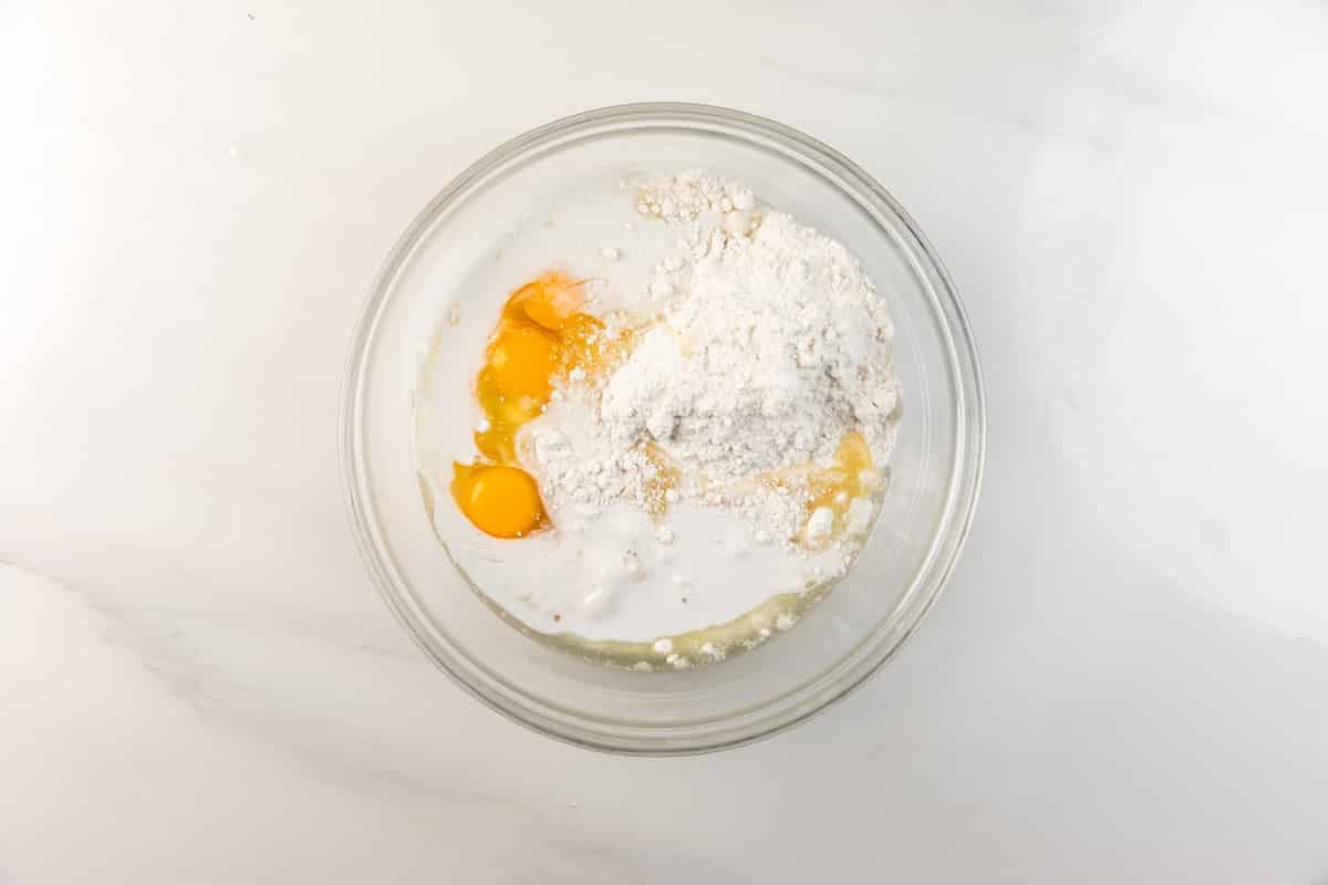 A glass bowl on a white surface containing cracked eggs, flour, and milk, ready to be mixed, viewed from above.