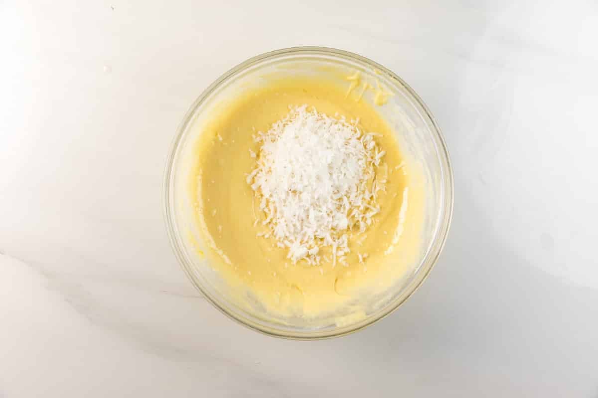 A glass bowl filled with yellow cake batter sits on a white surface. A mound of shredded coconut is piled in the center of the batter, ready to be mixed in.
