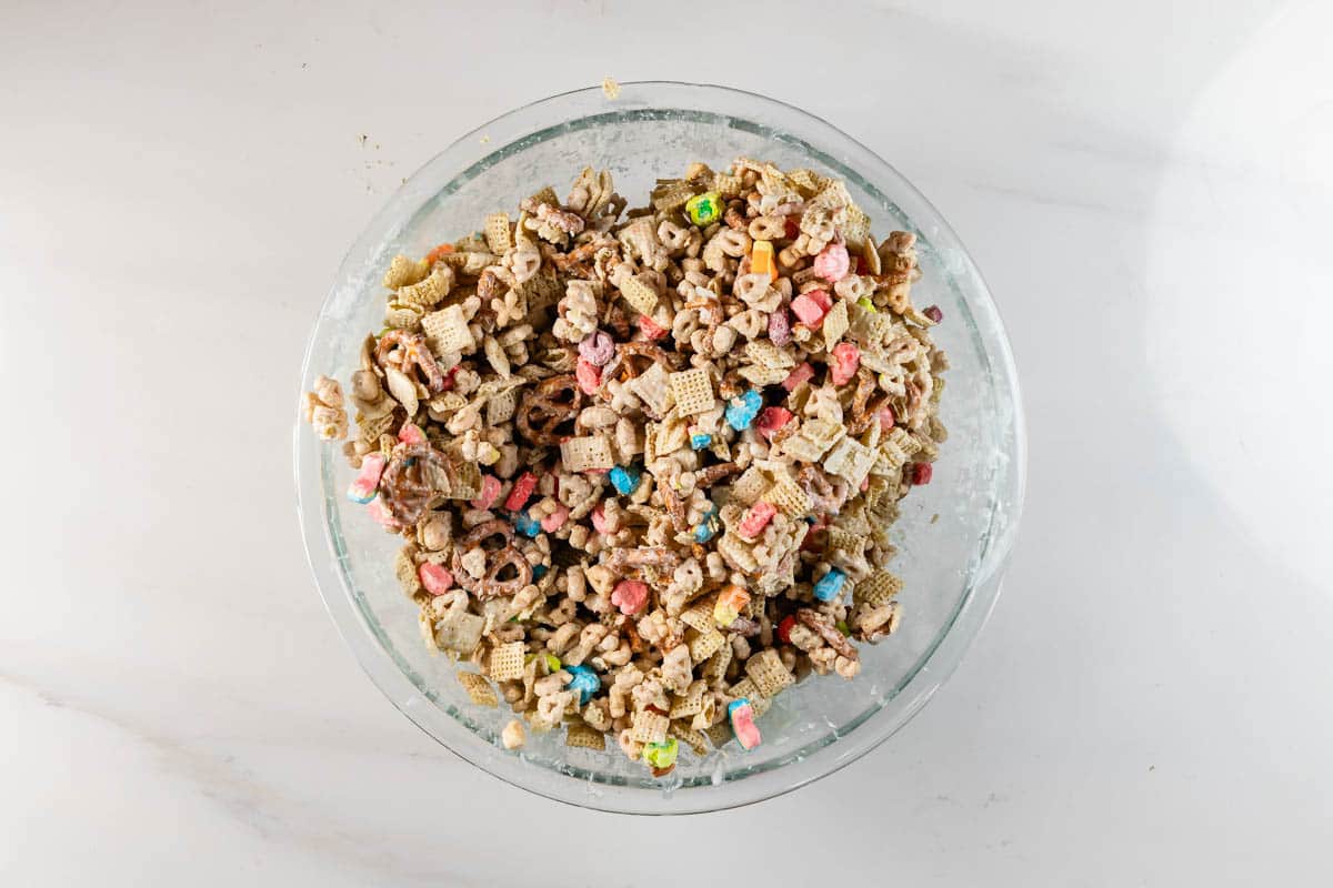 A glass bowl filled with a mixed cereal snack, including colorful marshmallows, pretzels, and various types of cereal pieces, on a white surface.