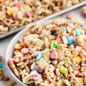 A white bowl filled with a colorful snack mix of cereal squares, pretzel sticks, mini marshmallows, and coated clusters; a tray with more of the mix is in the background.