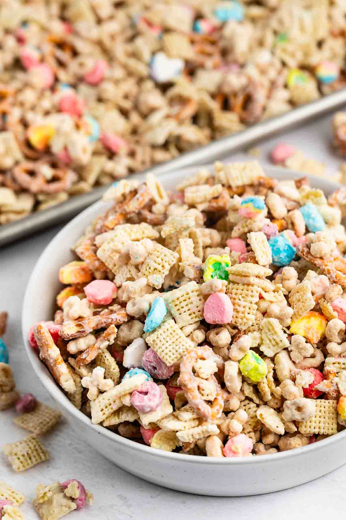 A white bowl filled with a colorful snack mix of cereal squares, pretzel sticks, mini marshmallows, and coated clusters; a tray with more of the mix is in the background.