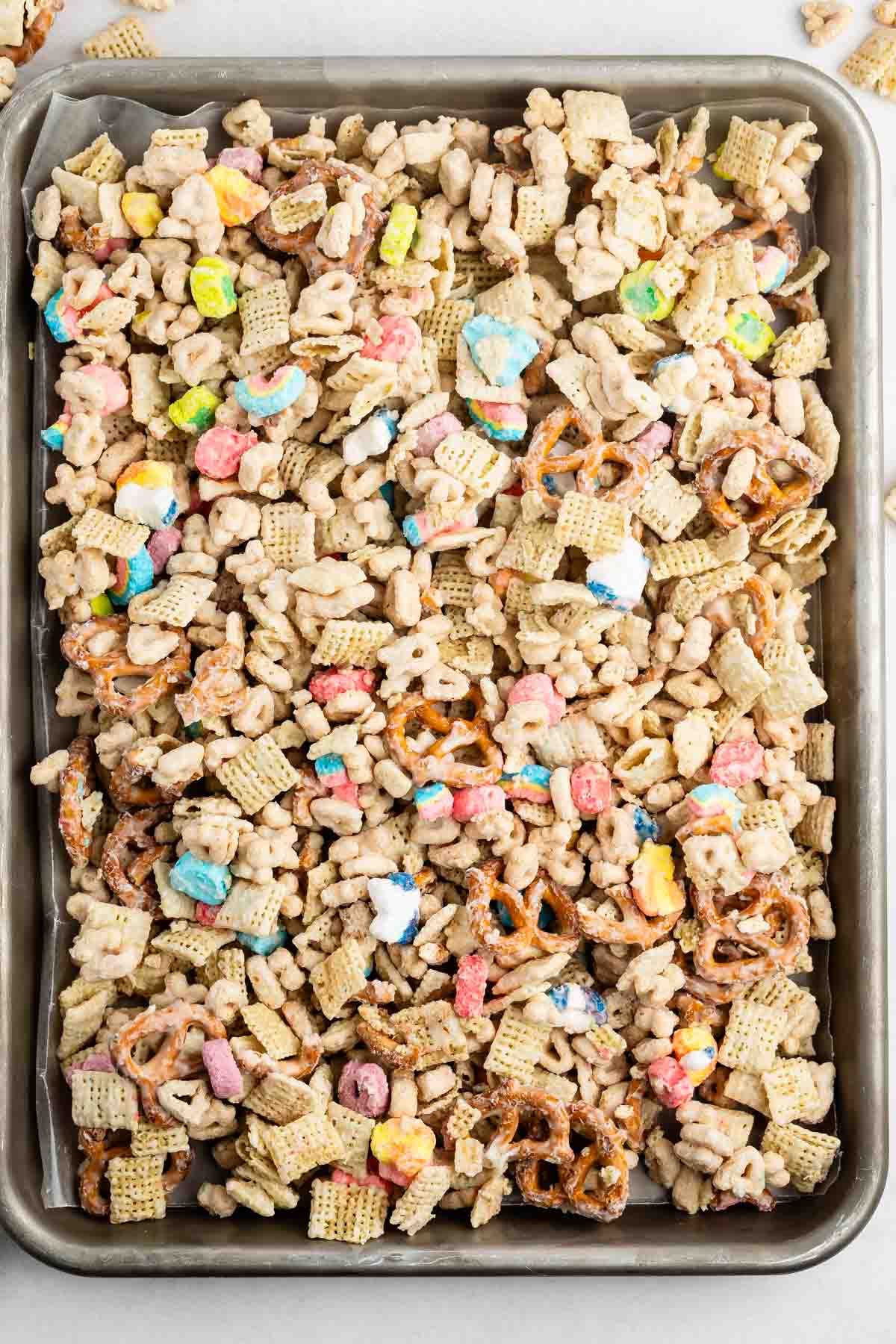 A baking sheet filled with a colorful snack mix, including pretzels, Chex cereal, and marshmallow cereal pieces, all coated in a white glaze or melted coating.