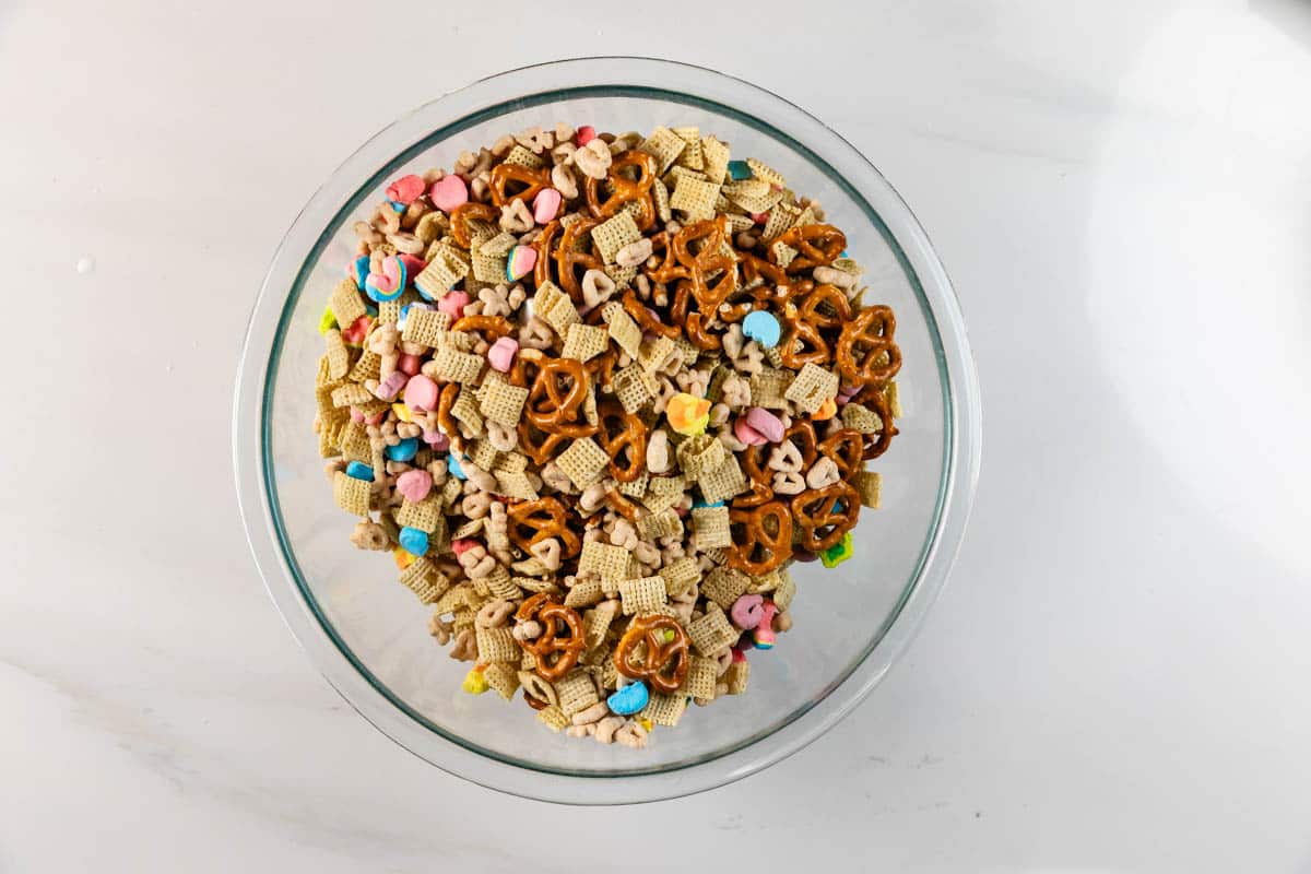 A glass bowl filled with a snack mix containing Chex cereal, pretzels, colorful marshmallows, and small oat cereal pieces, set on a white surface.