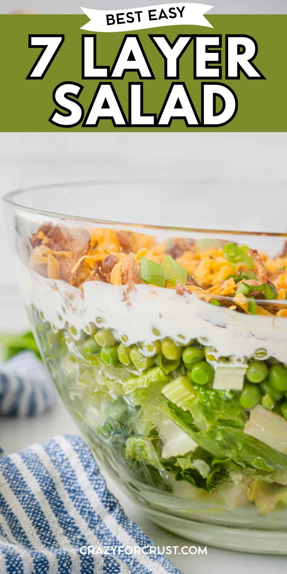 A clear glass bowl shows distinct layers of a 7-layer salad, including lettuce, peas, green onions, cheese, bacon, and creamy dressing. A striped blue and white towel sits beside the bowl. Text reads Best Easy 7 Layer Salad.