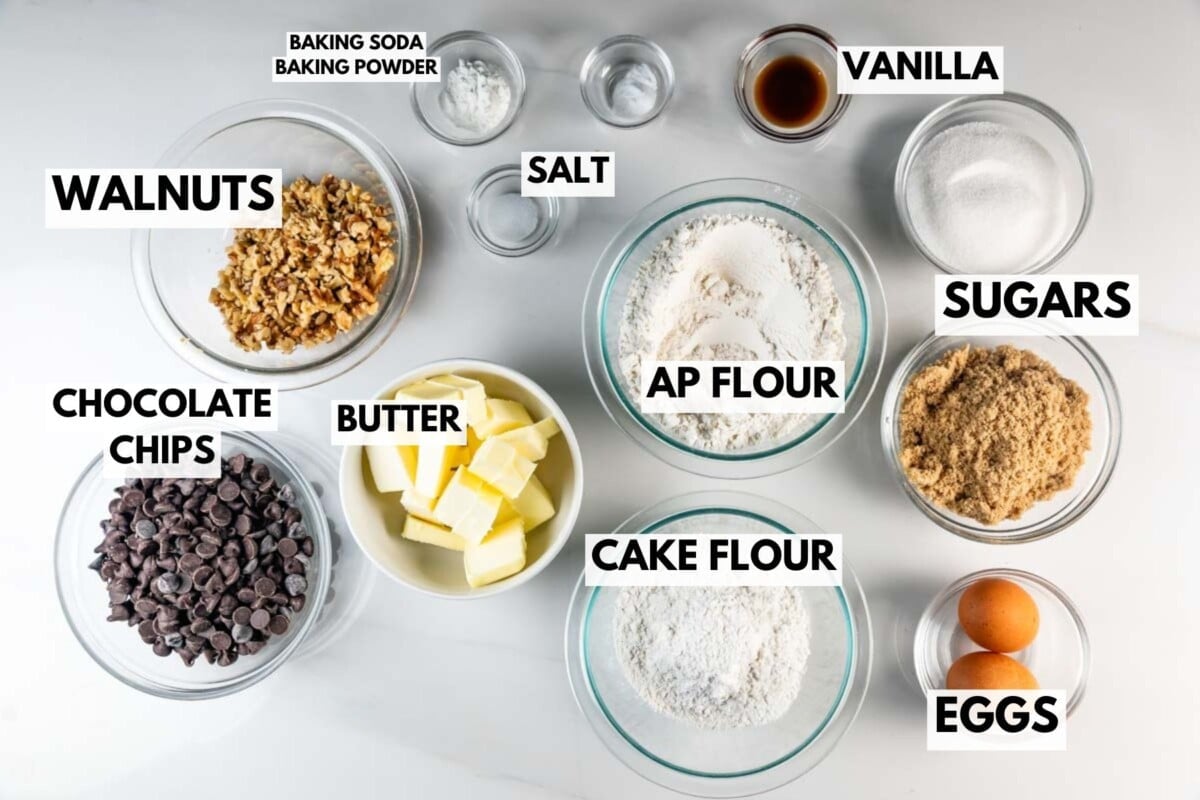 Bowls on a white surface containing baking ingredients labeled as walnuts, chocolate chips, butter, cake flour, all-purpose flour, salt, sugars, eggs, vanilla, baking soda, and baking powder.