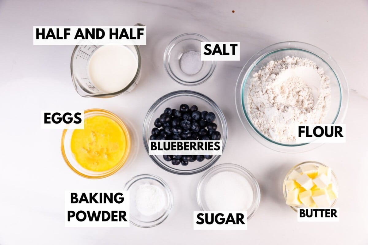 Top-down view of ingredients in bowls on a white surface, labeled: half and half, salt, flour, eggs, blueberries, baking powder, sugar, and butter, arranged neatly for baking.