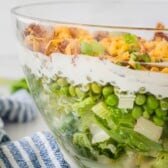 A clear glass bowl filled with layered salad, including lettuce, green peas, a creamy dressing, shredded cheese, bacon bits, and green onions, sitting next to a blue and white striped cloth.