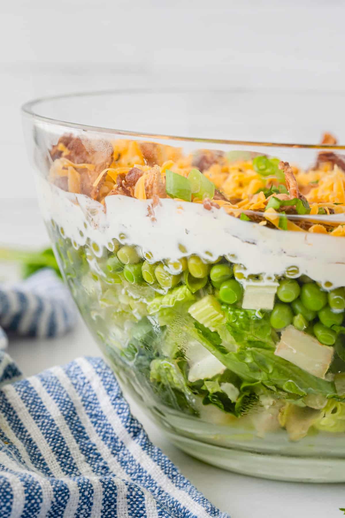 A clear glass bowl filled with layered salad, including lettuce, green peas, a creamy dressing, shredded cheese, bacon bits, and green onions, sitting next to a blue and white striped cloth.