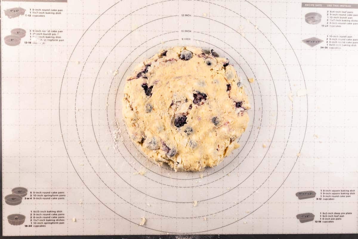 A round ball of dough with visible berries is placed on a pastry mat printed with concentric circles and measurements for rolling out dough to specific sizes.