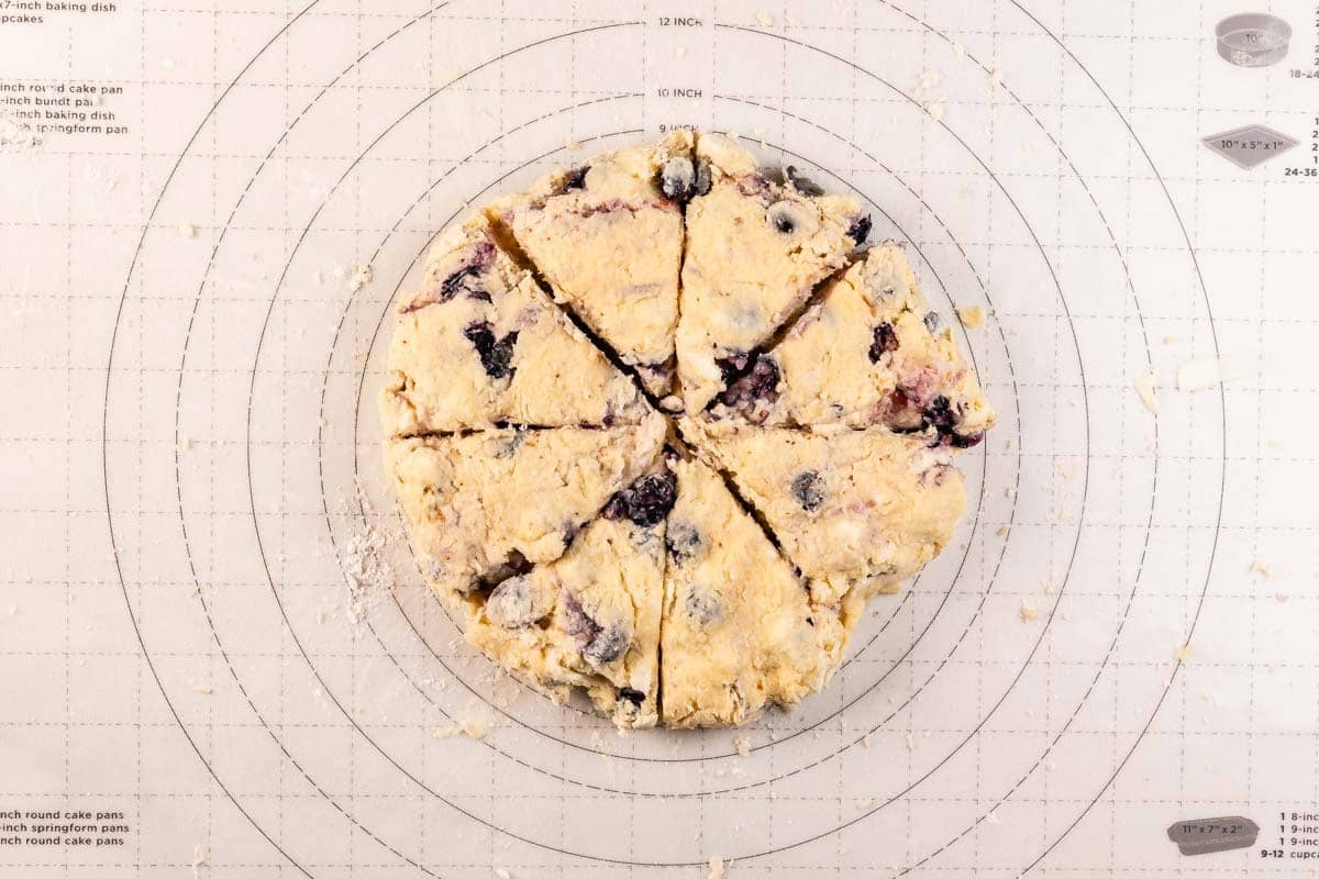A round disk of unbaked scone dough with blueberries, cut into eight wedges, sits on a pastry mat marked with measurement guides.