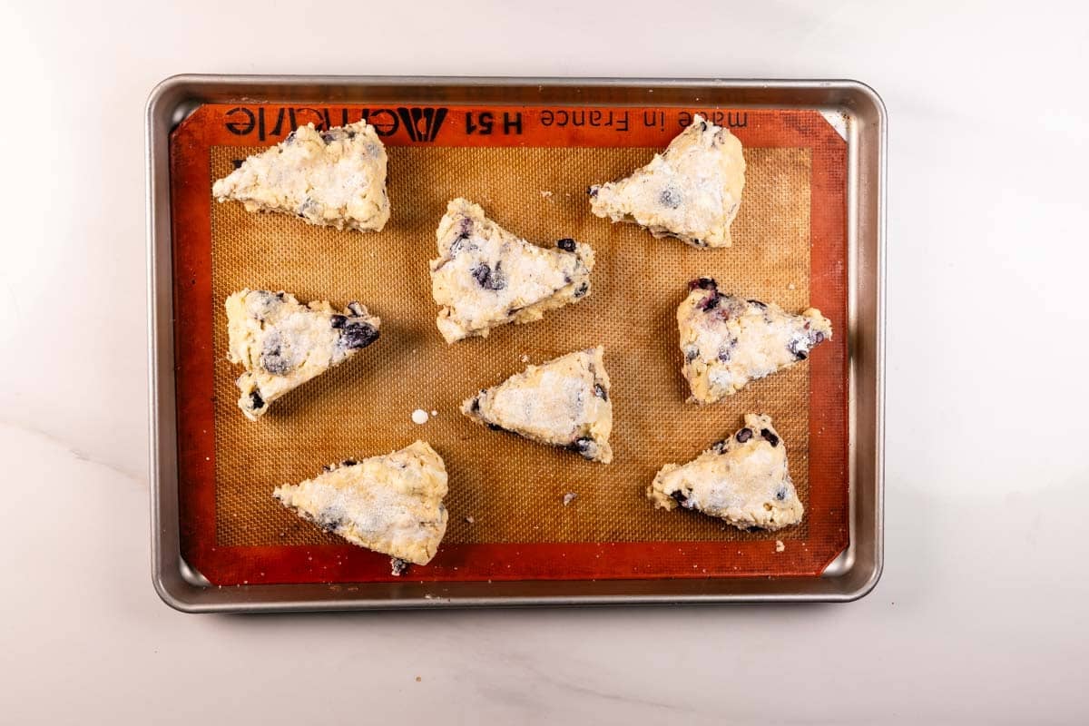 Eight unbaked, triangular scones with visible blueberries on a parchment-lined baking sheet, ready to be baked. The scones are lightly dusted with flour and arranged with some space between each.