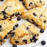 Golden-brown blueberry scones are arranged on a white plate, with a few fresh blueberries scattered around. The scones have a crumbly texture and visible blueberry pieces baked inside.