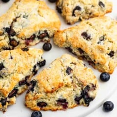 Golden-brown blueberry scones are arranged on a white plate, with a few fresh blueberries scattered around. The scones have a crumbly texture and visible blueberry pieces baked inside.
