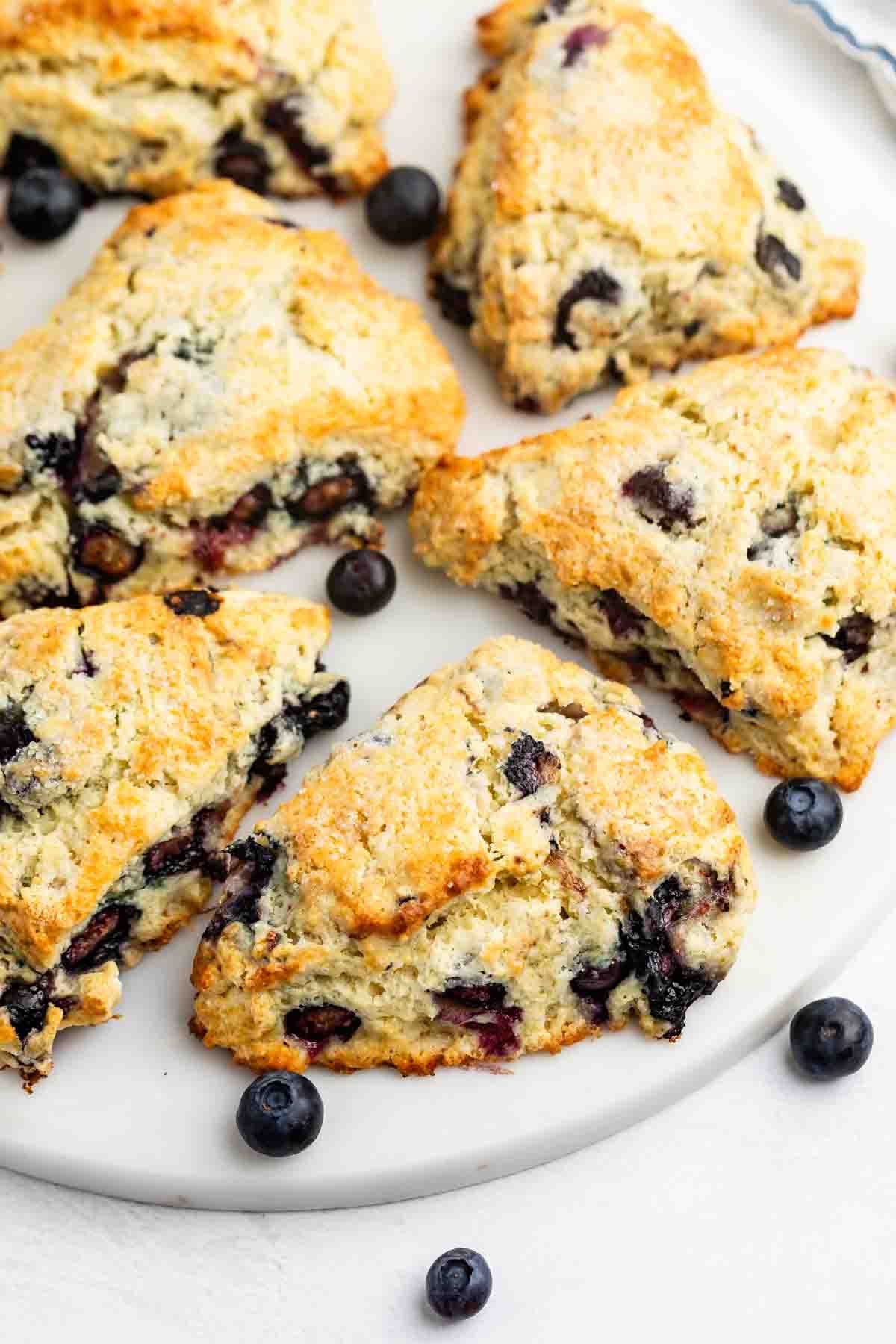 Golden-brown blueberry scones are arranged on a white plate, with a few fresh blueberries scattered around. The scones have a crumbly texture and visible blueberry pieces baked inside.