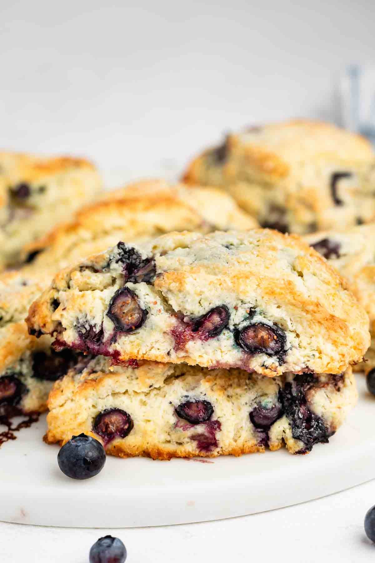 Close-up of a halved blueberry scone with visible whole blueberries, placed on a white surface with more scones and a few loose blueberries in the background. The scone looks freshly baked and crumbly.