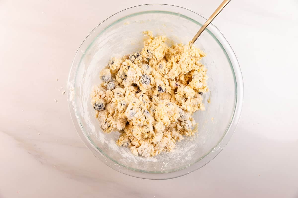 A glass mixing bowl filled with raw scone dough containing blueberries. A metal spoon is stuck into the thick, partially mixed dough on a white countertop.