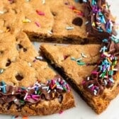 A decadent chocolate chip cookie cake topped with rich chocolate frosting and colorful sprinkles along the edge, featuring one triangular slice slightly pulled out to reveal the soft, gooey center.