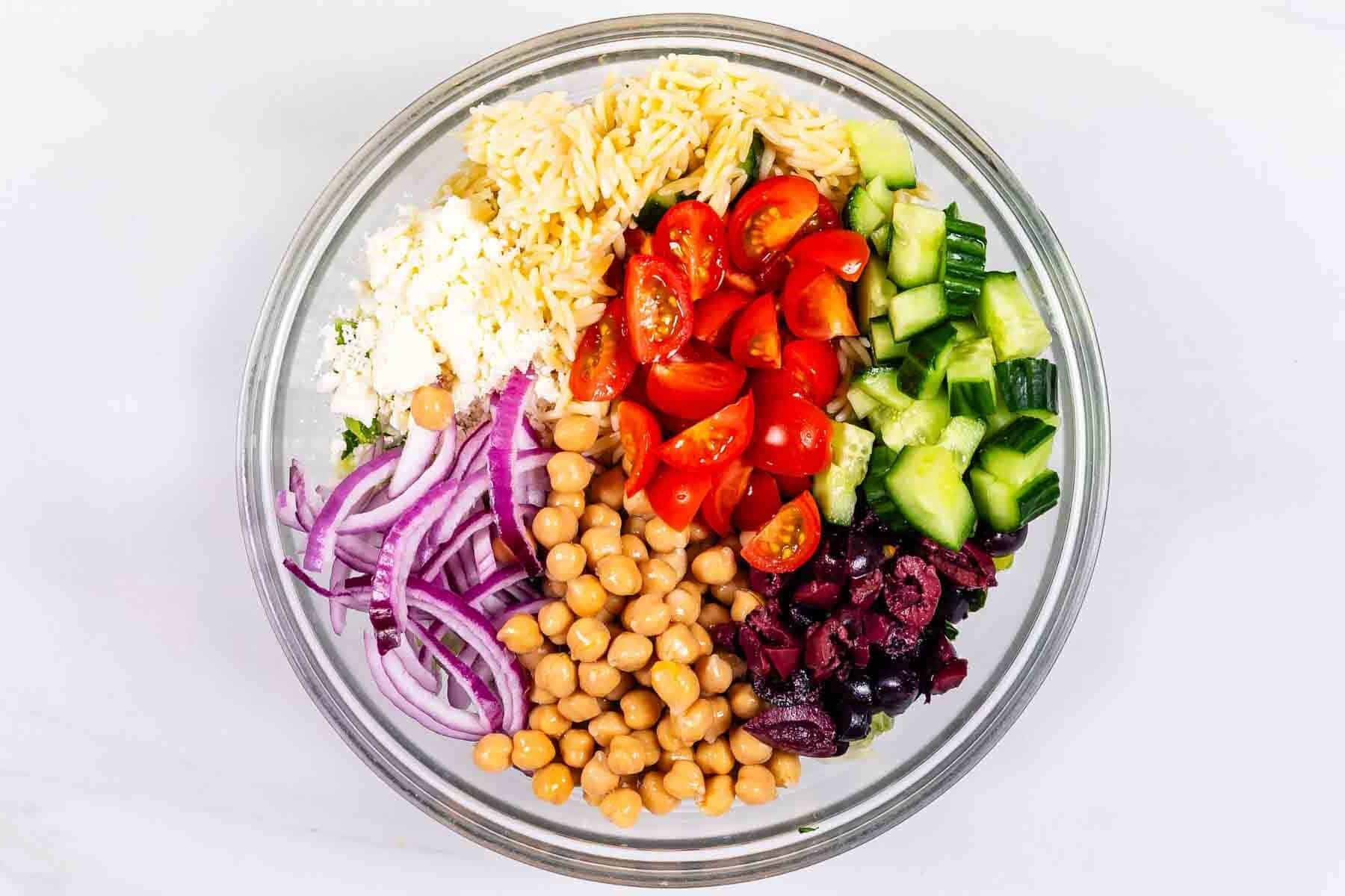 A glass bowl filled with chopped cucumber, cherry tomatoes, kalamata olives, chickpeas, sliced red onion, orzo pasta, and crumbled feta cheese, arranged in separate sections on a white background.