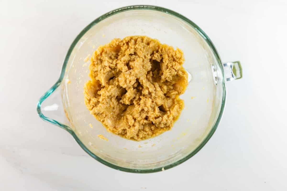 A glass mixing bowl on a white surface contains a thick, yellowish cookie dough mixture.