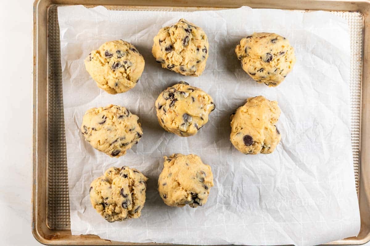 Seven large scoops of chocolate chip cookie dough are spaced apart on a parchment-lined baking sheet, ready to be baked.