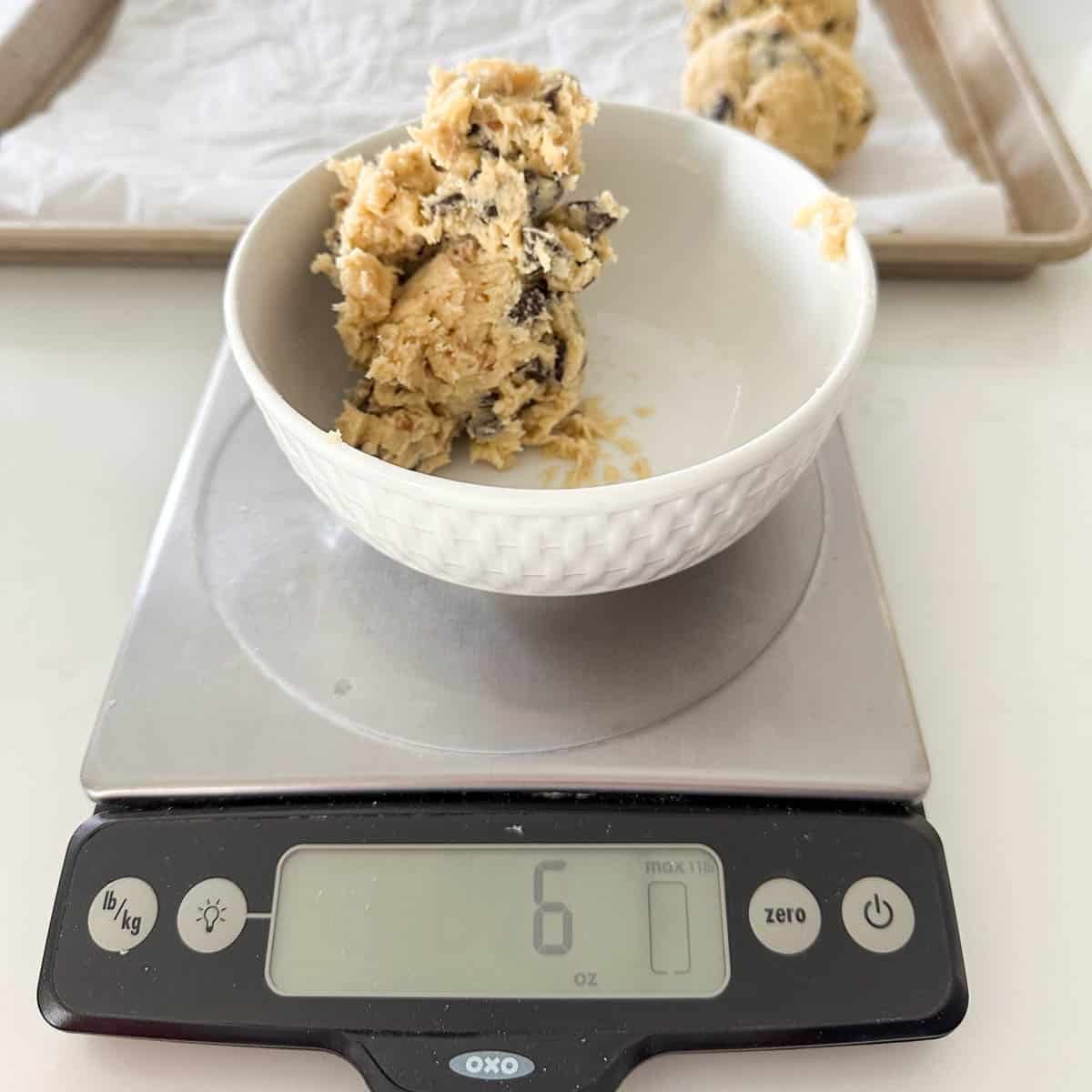 A white bowl with chocolate chip cookie dough sits on a digital kitchen scale displaying 6 ounces. In the background, cookie dough balls rest on a parchment-lined baking sheet.