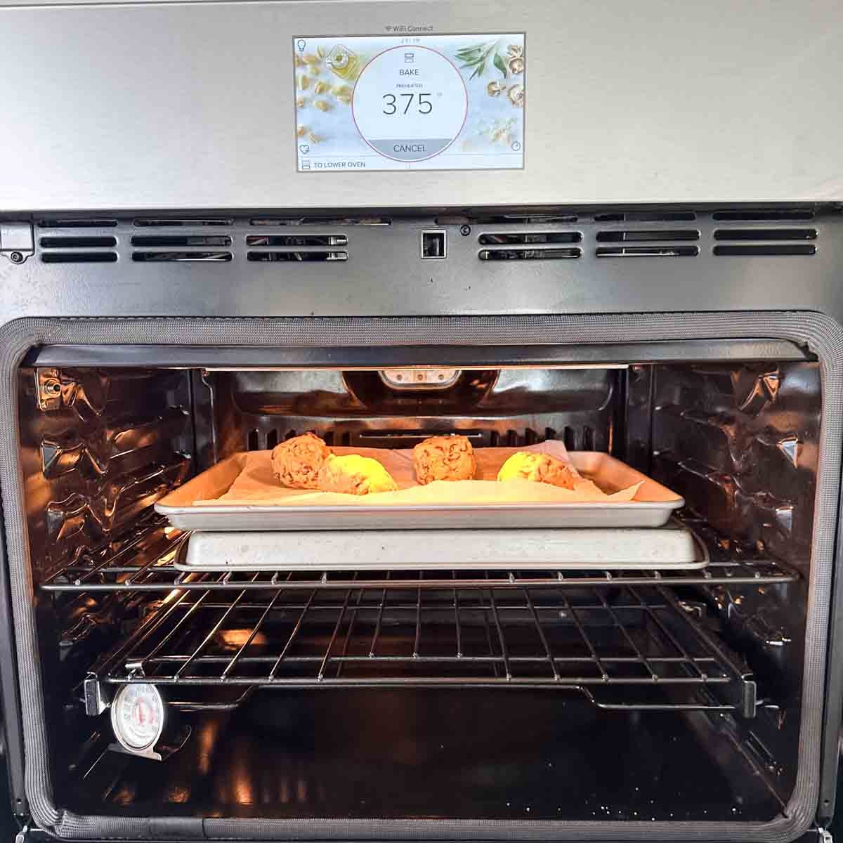 A tray of cookie dough balls baking in an oven, with an oven thermometer on the rack and the digital display above showing a temperature of 375°F.