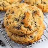 Two thick chocolate chip cookies are stacked on a cooling rack, with more cookies and scattered chocolate chips in the background. The cookies are golden brown with visible chocolate chips.