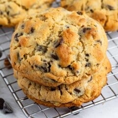 Two thick chocolate chip cookies are stacked on a cooling rack, with more cookies and scattered chocolate chips in the background. The cookies are golden brown with visible chocolate chips.