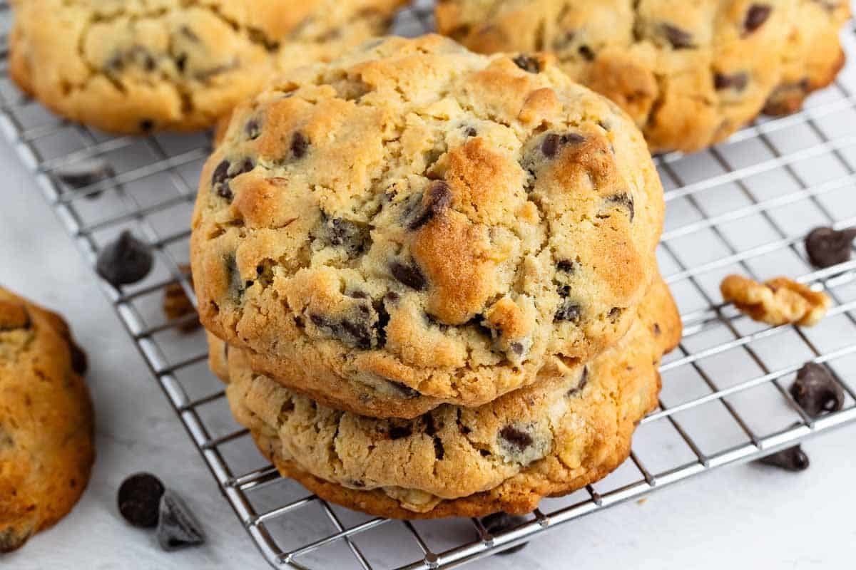 Two thick chocolate chip cookies are stacked on a cooling rack, with more cookies and scattered chocolate chips in the background. The cookies are golden brown with visible chocolate chips.